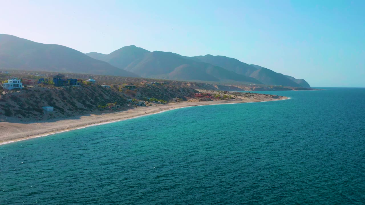 Aerial view one second in at El Sargento La Ventana, Mexico, showing the shoreline arc more centered, teal water below, mountain ridges in the distance, and no visible people.