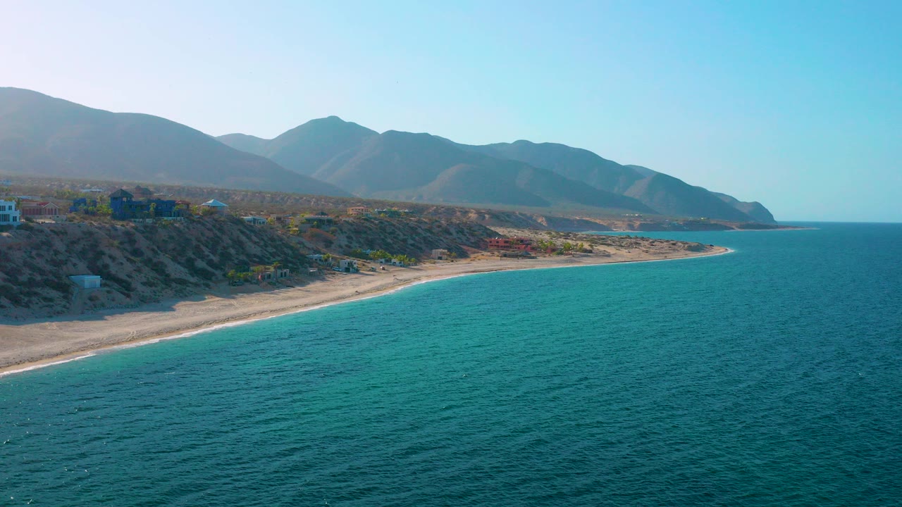 Elevated 4K view of El Sargento La Ventana, Mexico with turquoise water in front, a sandy shoreline below the bluff, hazy mountains behind, and no visible people.
