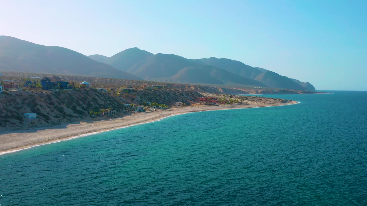Aerial view over El Sargento La Ventana, Mexico with more open ocean in the foreground, a thin pale beach line at left, layered mountain slopes in the distance, and no visible people.