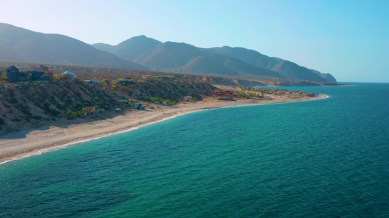 Aerial coastal frame from El Sargento La Ventana, Mexico, with more open sea on the right, a narrow beach tracing the land, dry hills above it, distant mountains, and no visible people.