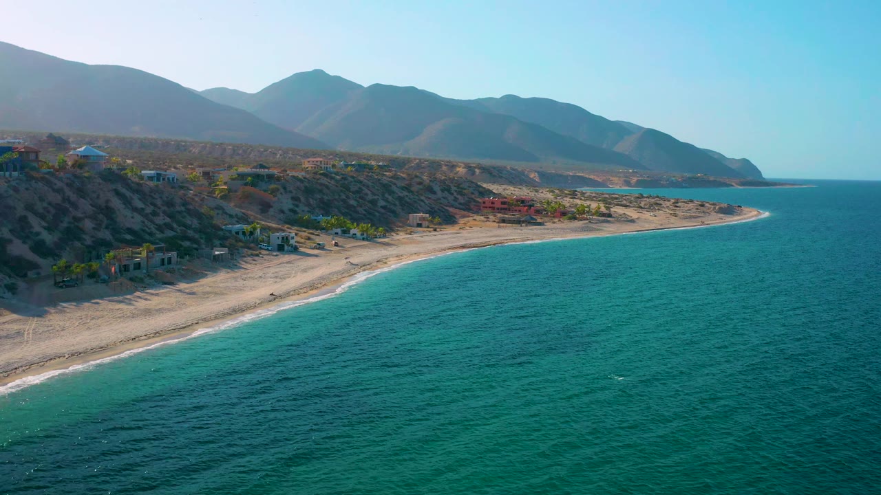 Wide aerial coastline in El Sargento La Ventana, Mexico, showing blue-green water, a sandy shoreline, scattered coastal buildings, mountains, and no visible people.