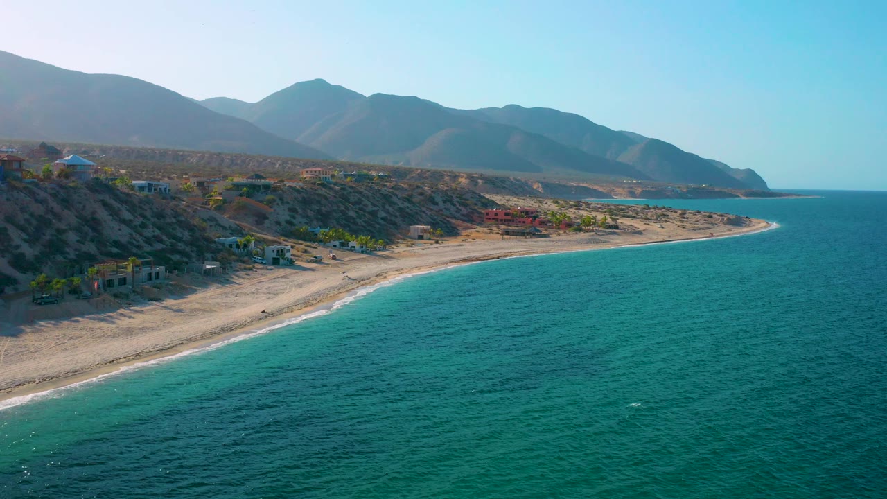 Aerial coastal frame from El Sargento La Ventana, Mexico, with broad turquoise ocean foreground, a pale beach curving toward the right, mountain ridges, and no visible people.