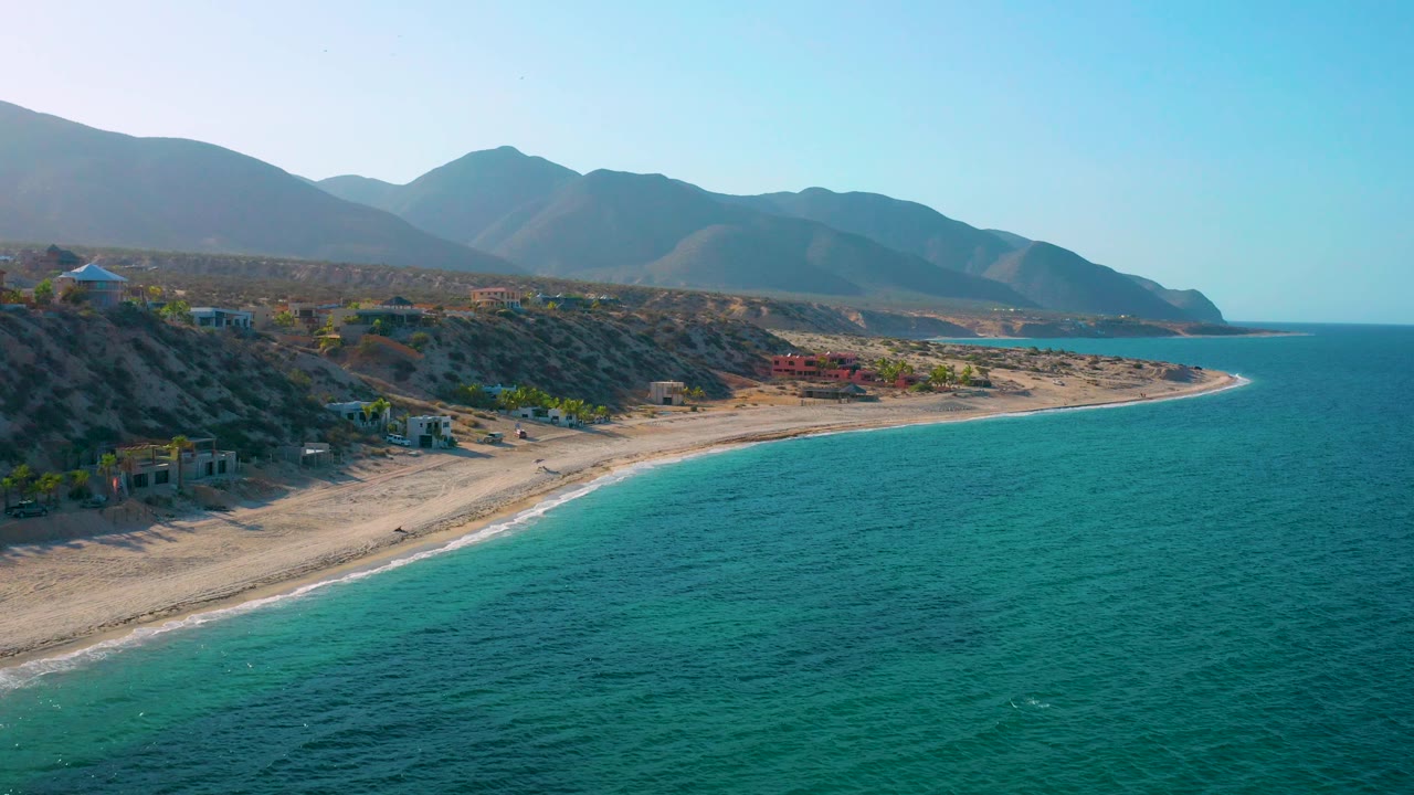 Coastal aerial view with hillside buildings, sandy beach, turquoise water, and mountains.