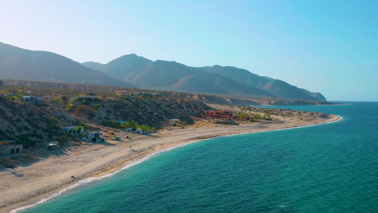 Aerial opening view over El Sargento La Ventana, Mexico, with turquoise water on the right, a pale curved beach below, desert bluffs inland, mountains on the horizon, and no visible people.