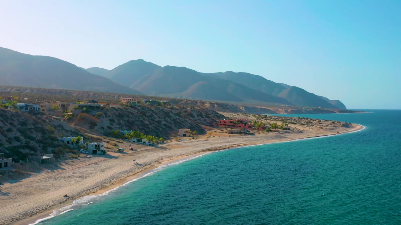 Drone view along the El Sargento La Ventana shoreline, with the sea taking more of the right frame, a narrow surf edge tracing the beach, low buildings inland, and no visible people.