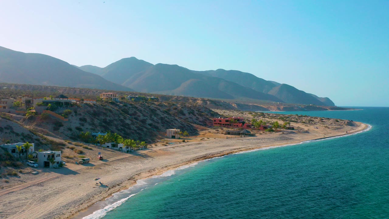 Opening high view of El Sargento La Ventana coastline with turquoise water at right, a curved sandy beach, dry hillside buildings, distant mountains, and no visible people.