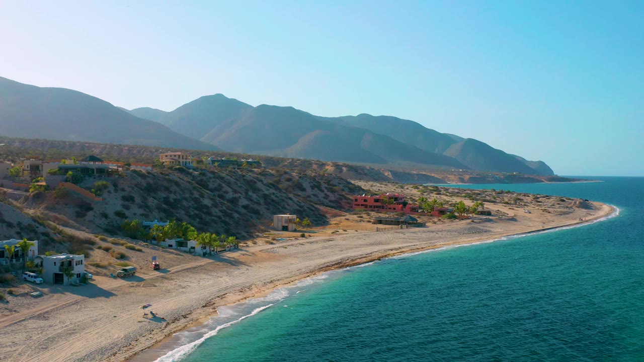 Wide aerial view of El Sargento La Ventana with open sea filling the right side, a pale beach bending toward the horizon, hillside structures, layered mountains, and no visible people.