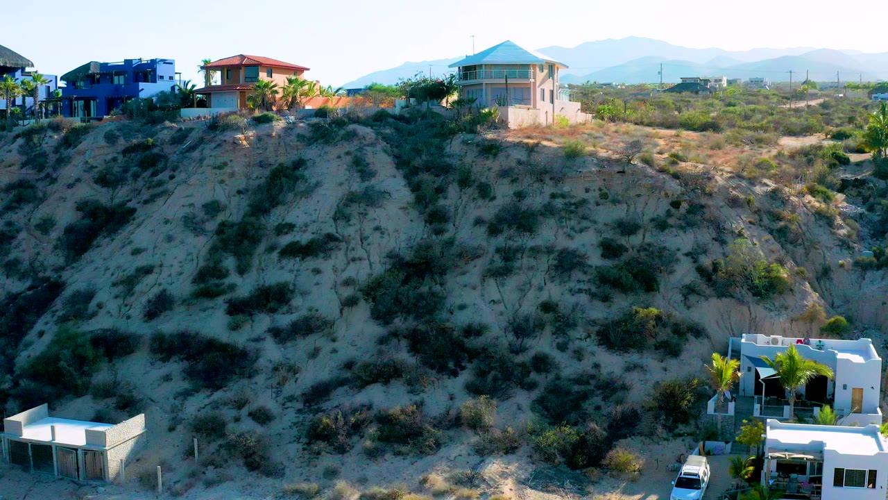 Wide elevated view in El Sargento La Ventana, Mexico showing blue, red-roofed, and pale-roofed houses above a brushy sandy hill with hazy mountains behind and no visible people.