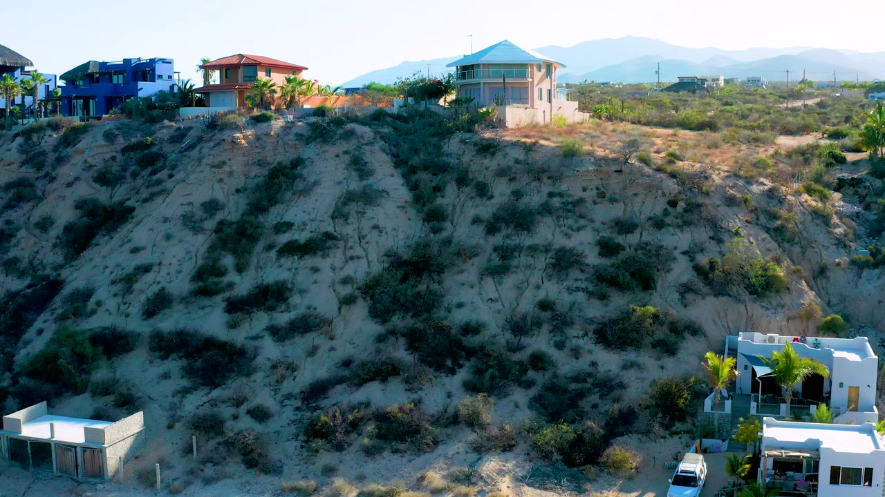 Hillside residential view in El Sargento La Ventana, Mexico with the blue house near the left ridge, a red-roofed house near center, a pale-roofed house to the right, scrub terrain below, and no visible people.