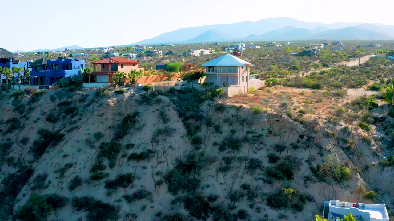 Aerial frame in El Sargento La Ventana, Mexico, holding on the blue and red-roofed houses near the cliff edge, with desert scrub, unpaved roads, distant homes, and no visible people.