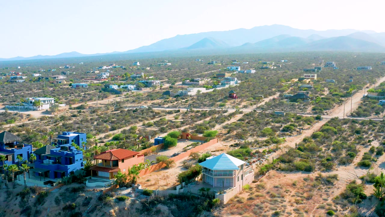 Aerial frame from El Sargento La Ventana, Mexico with blue foreground homes, a red-roofed house, branching dirt roads, open scrub lots, hazy mountain ridges, and no visible people.