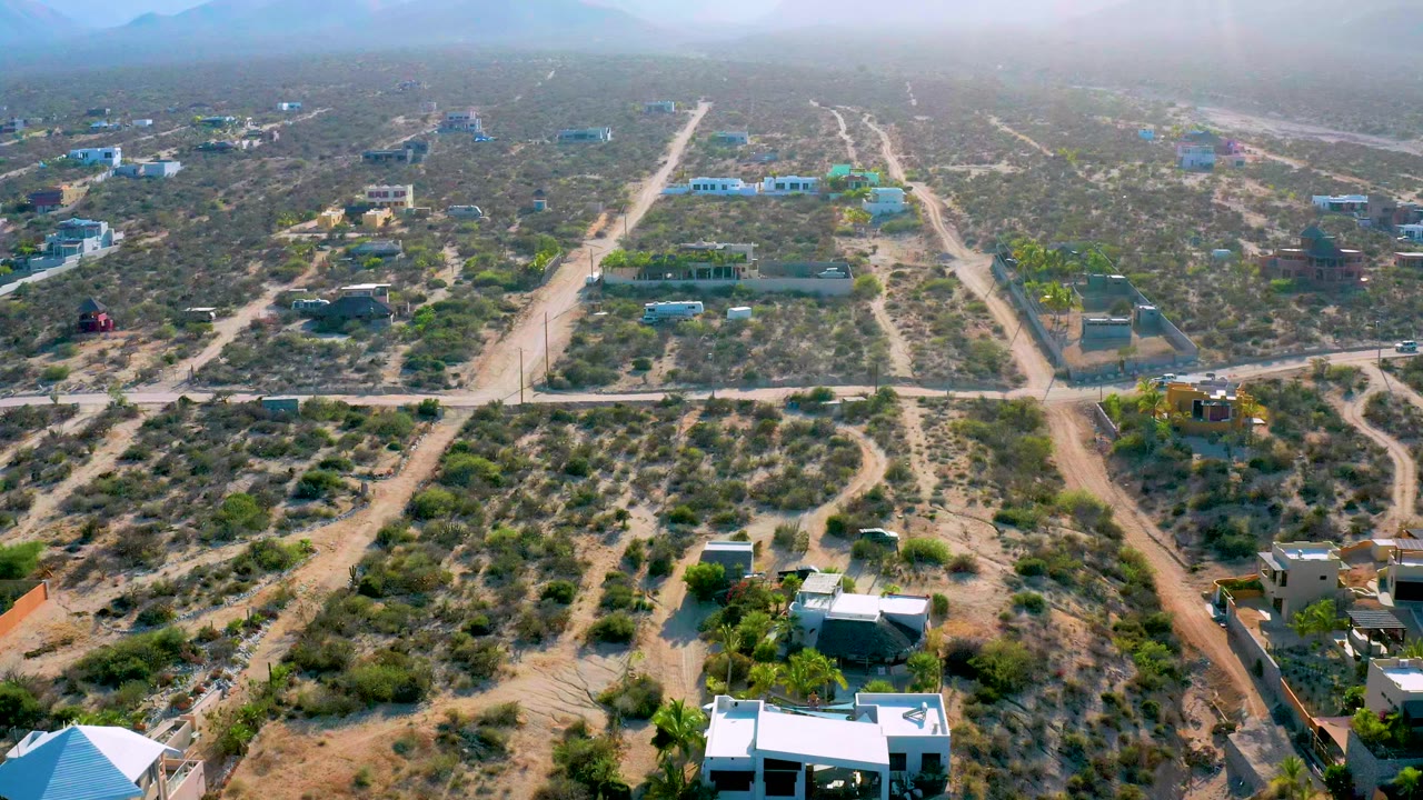 High aerial frame in El Sargento La Ventana showing unpaved roads crossing dry scrubland, scattered homes, walled lots, and a hazy mountain horizon, with no visible people.