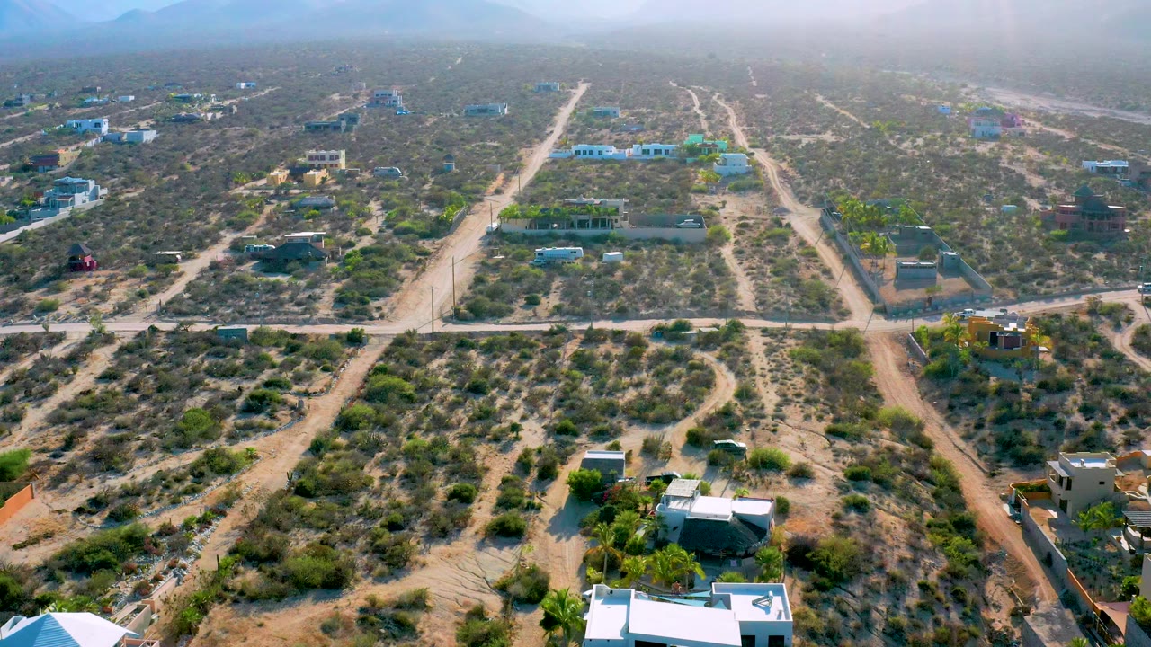 Wide aerial view of El Sargento La Ventana with pale dirt roads, desert vegetation, small buildings, green tree clusters, and soft haze over the mountains, with no visible people.