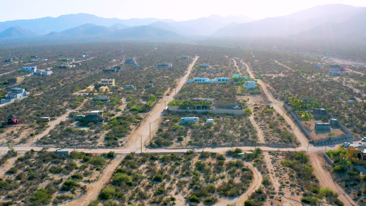 Opening aerial frame over El Sargento La Ventana, Mexico, showing scrubby desert lots, pale dirt roads, separated buildings, hazy mountains, and no visible people.