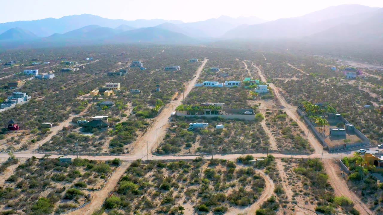 Second frame over El Sargento La Ventana, Mexico, with a central dirt road, low green bushes, small separated buildings, distant blue mountains, and no visible people.
