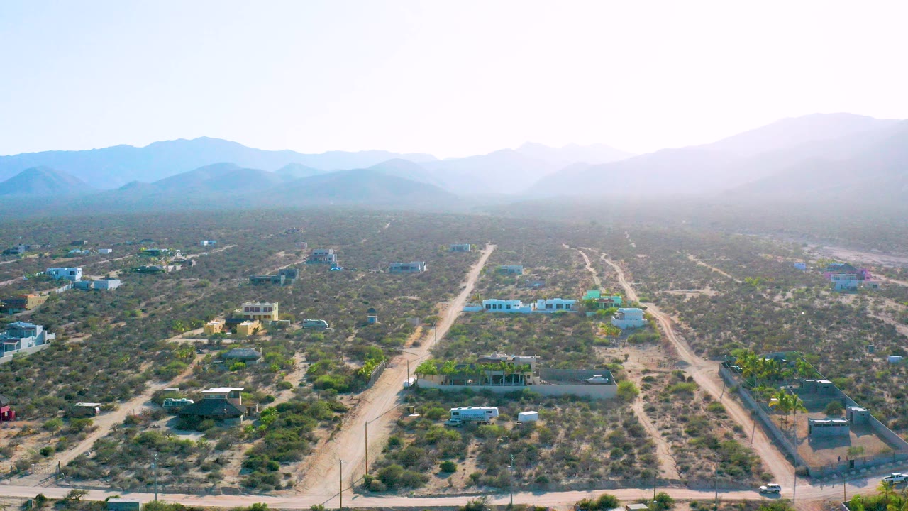 Opening aerial frame over El Sargento La Ventana, Mexico, with intersecting dirt roads, scattered low houses, arid vegetation, hazy mountains, bright sky, and no visible people.