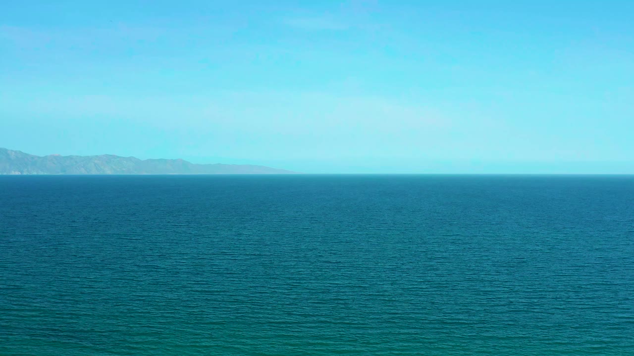Wide aerial view of blue ocean at El Sargento La Ventana, Mexico, with distant low land across the horizon and no visible people.