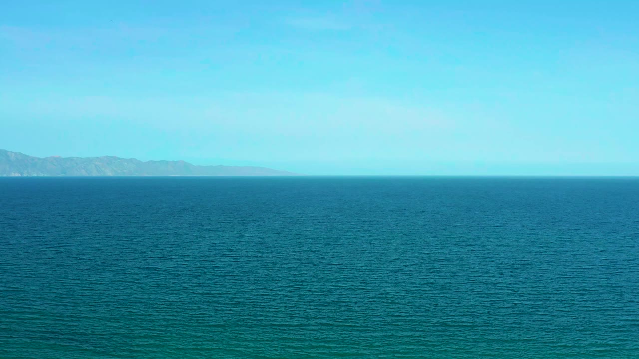 Aerial ocean frame from El Sargento La Ventana, Mexico, showing teal water, low distant mountains near the horizon, and no visible people.
