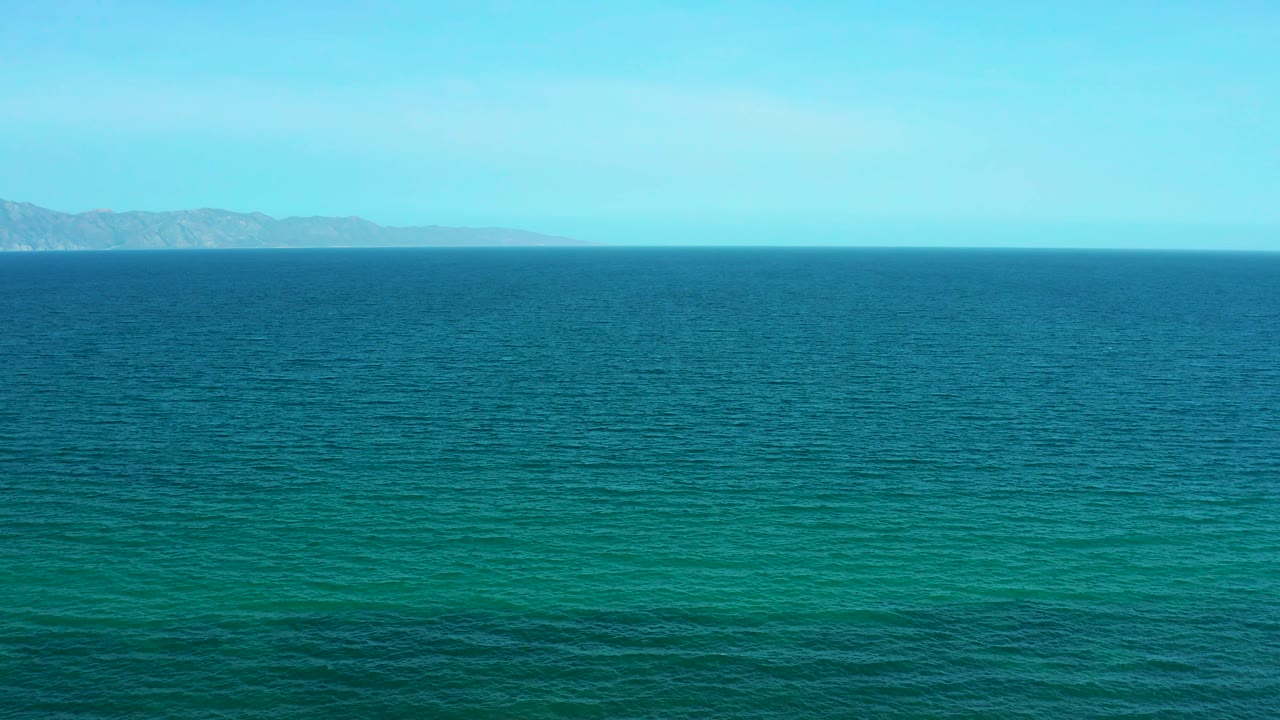 A wide view of turquoise and deeper blue ocean shows distant mountains softened by haze near El Sargento La Ventana, Mexico, with no visible people.
