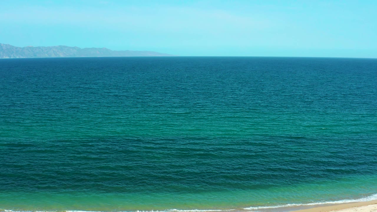 Turquoise water darkens into deeper blue offshore as small waves reach the sand in El Sargento La Ventana, Mexico, with no visible people.