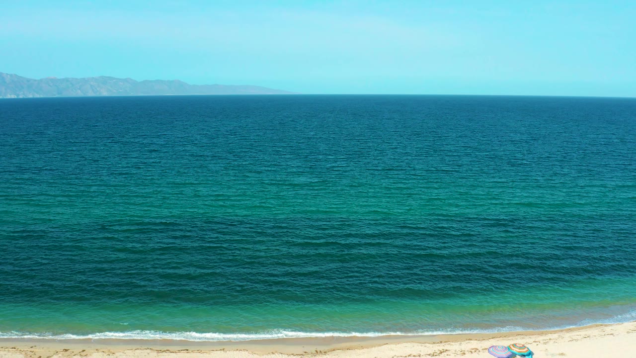 Blue ocean stretches toward distant land while a pale strip of sand and low white waves remain visible at the bottom in El Sargento La Ventana, Mexico, with no visible people.