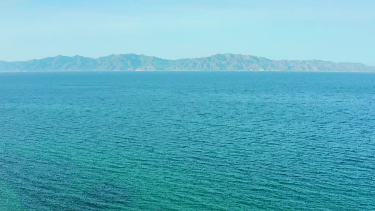 Rippling ocean water in El Sargento La Ventana, Mexico, sits below distant arid-looking mountains and a pale blue sky, with no visible people.