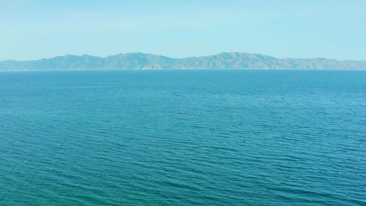 A wide view of turquoise ocean in El Sargento La Ventana, Mexico, with low surface ripples, a distant mountain range, pale blue sky, and no visible people.