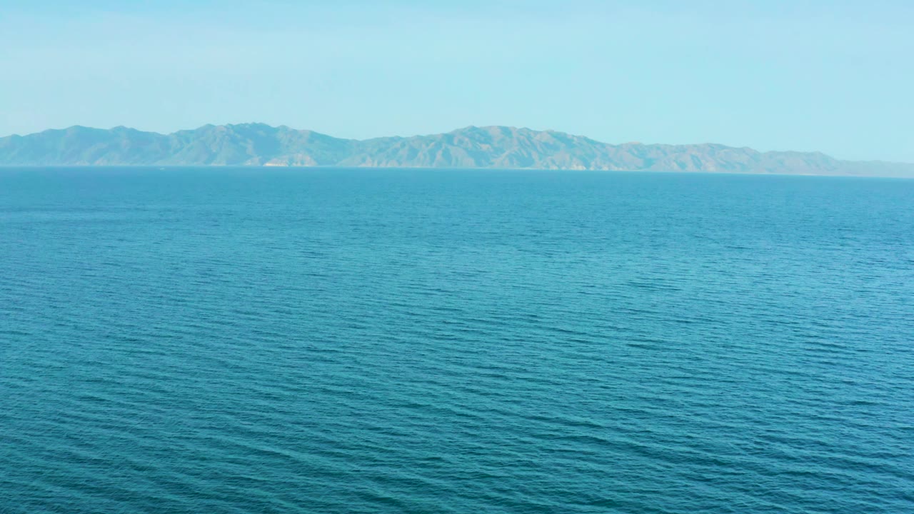Second frame of El Sargento La Ventana, Mexico, with broad blue water in the foreground and rugged tan-gray mountains stretching across the background, with no visible people.