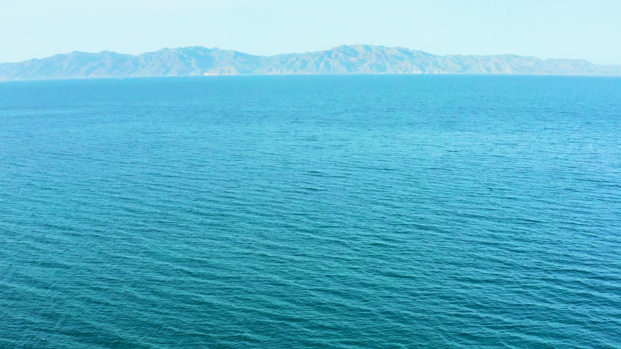 Wide view over rippling turquoise water toward distant mountains in El Sargento La Ventana, Mexico, with pale sky and no visible people.