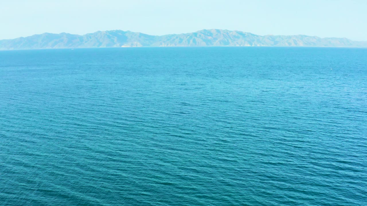 A broad turquoise sea surface shows fine wave lines leading to distant mountains and pale sky in El Sargento La Ventana, Mexico, with no visible people.