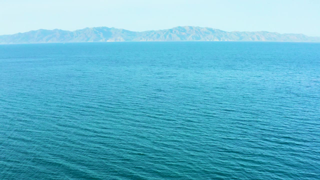 The ocean appears slightly lighter blue as small ripples spread across the frame toward a long mountain horizon in El Sargento La Ventana, Mexico, with no visible people.