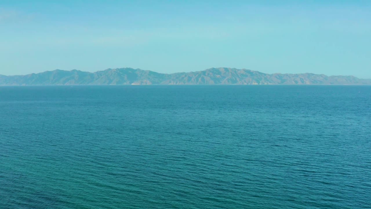 Frame at 0:02 in El Sargento La Ventana, Mexico, showing turquoise-blue ocean texture, pale clear sky, and distant dry hills along the horizon, with no visible people.