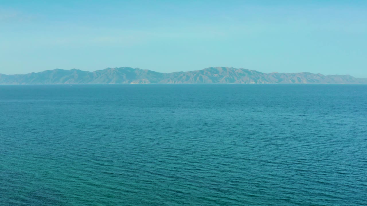 Mid-clip frame from El Sargento La Ventana, Mexico, where open blue sea occupies the lower frame and hazy mountains sit beneath a clear sky, with no visible people.