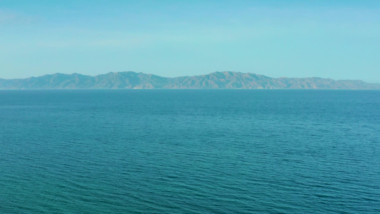 Wide view of aquamarine open water near El Sargento La Ventana, Mexico, with ripples in the foreground, distant low mountains on the horizon, hazy blue sky, and no visible people.
