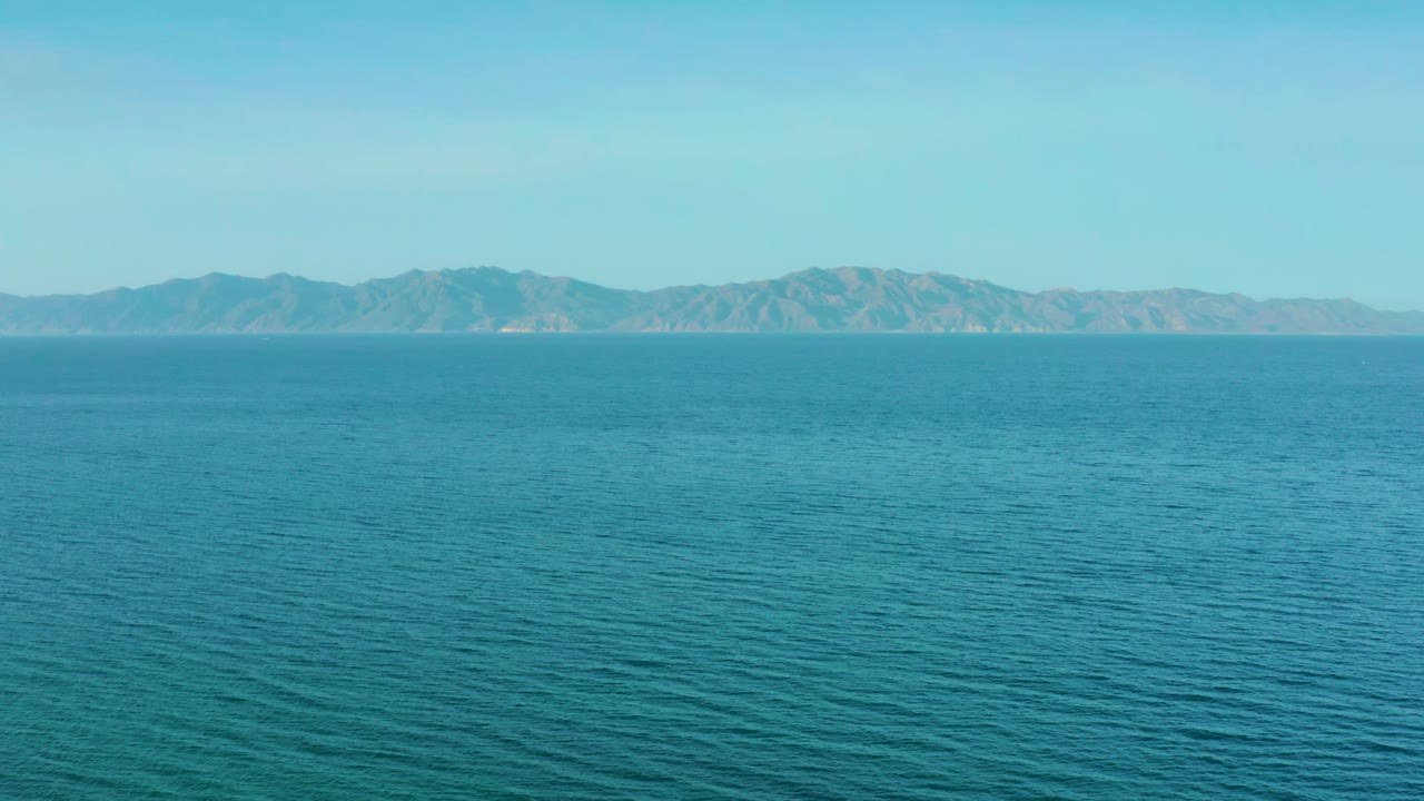 Aquamarine water fills most of the frame near El Sargento La Ventana, Mexico, with a broad band of distant mountains, pale haze, and no visible people.