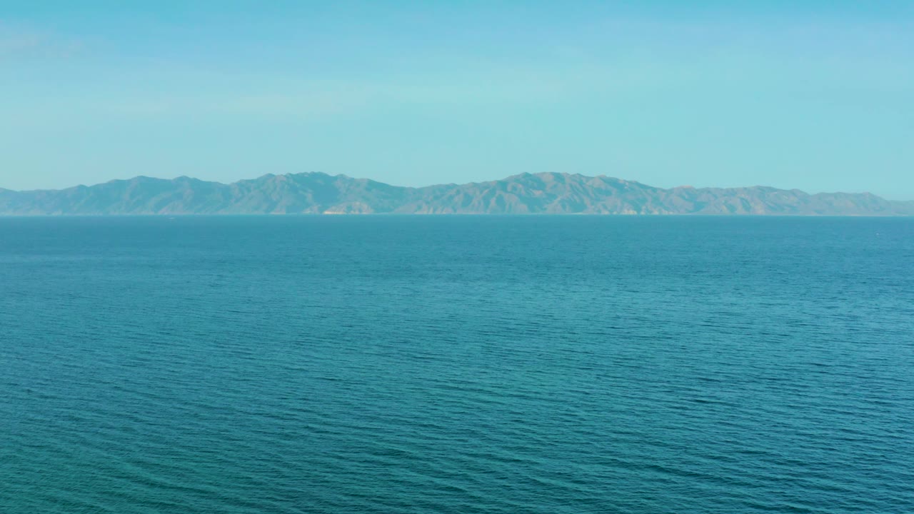 Mid-clip frame of blue-green sea near El Sargento La Ventana, Mexico, with a low mountain horizon, soft white haze above the land, and no visible people.
