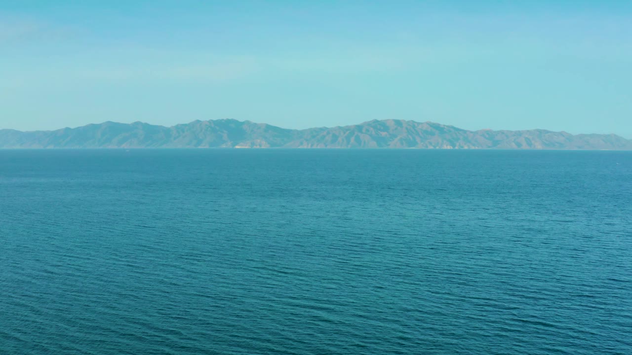 Blue sea with fine ripples leads toward a low tan mountain range under clear daylight near El Sargento La Ventana, Mexico, with no visible people.