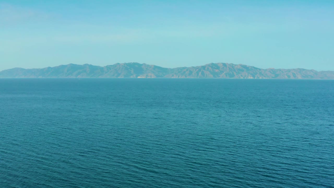 A broad expanse of blue water leads to a distant mountain chain under clear daylight near El Sargento La Ventana, Mexico, with no visible people.