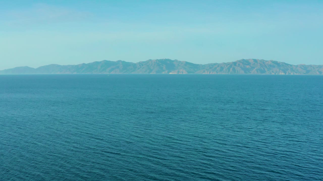 At 0:03, calm-looking ocean water fills the lower frame while blue-gray mountains remain distant under a clear sky in El Sargento La Ventana, with no visible people.