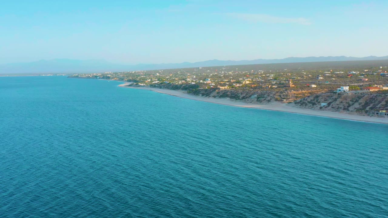 Opening aerial frame over blue water in El Sargento La Ventana, Mexico, with rippled ocean foreground, sandy beach, low coastal homes, distant mountains, and no visible people.