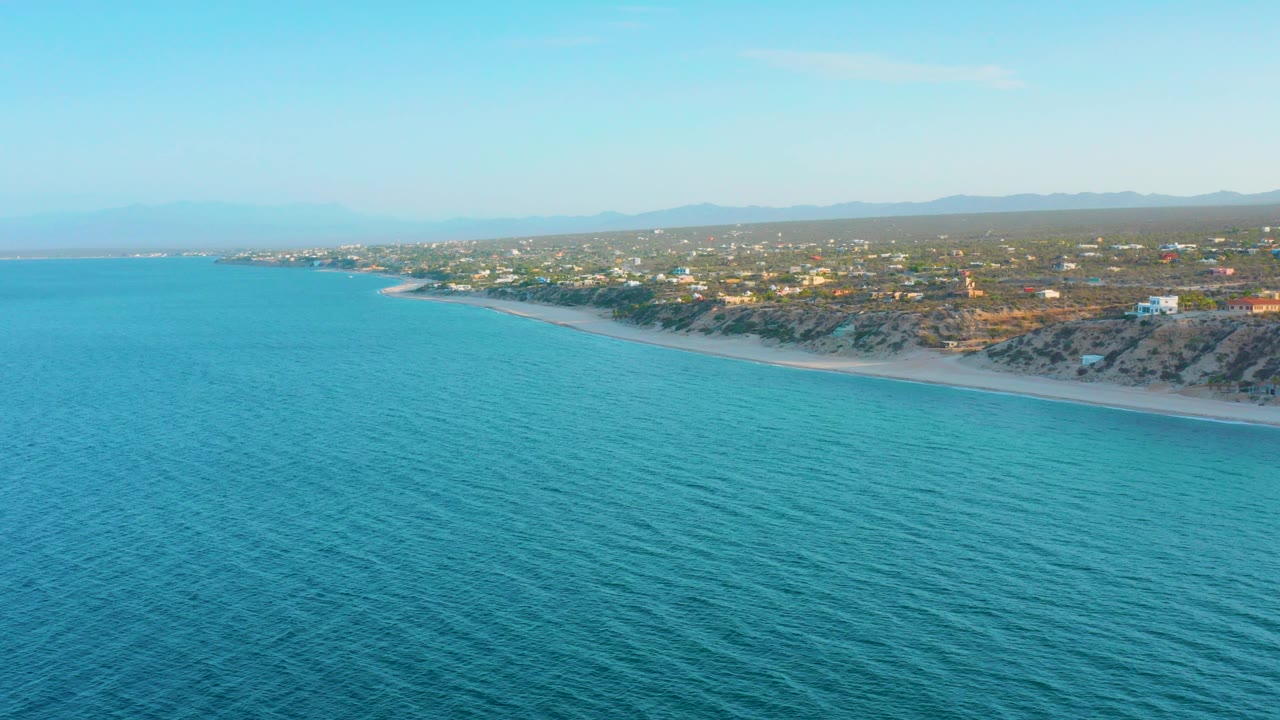 Opening aerial frame over turquoise water facing the El Sargento La Ventana coastline, with a thin sandy beach, scattered inland buildings, distant hills, and no visible people.