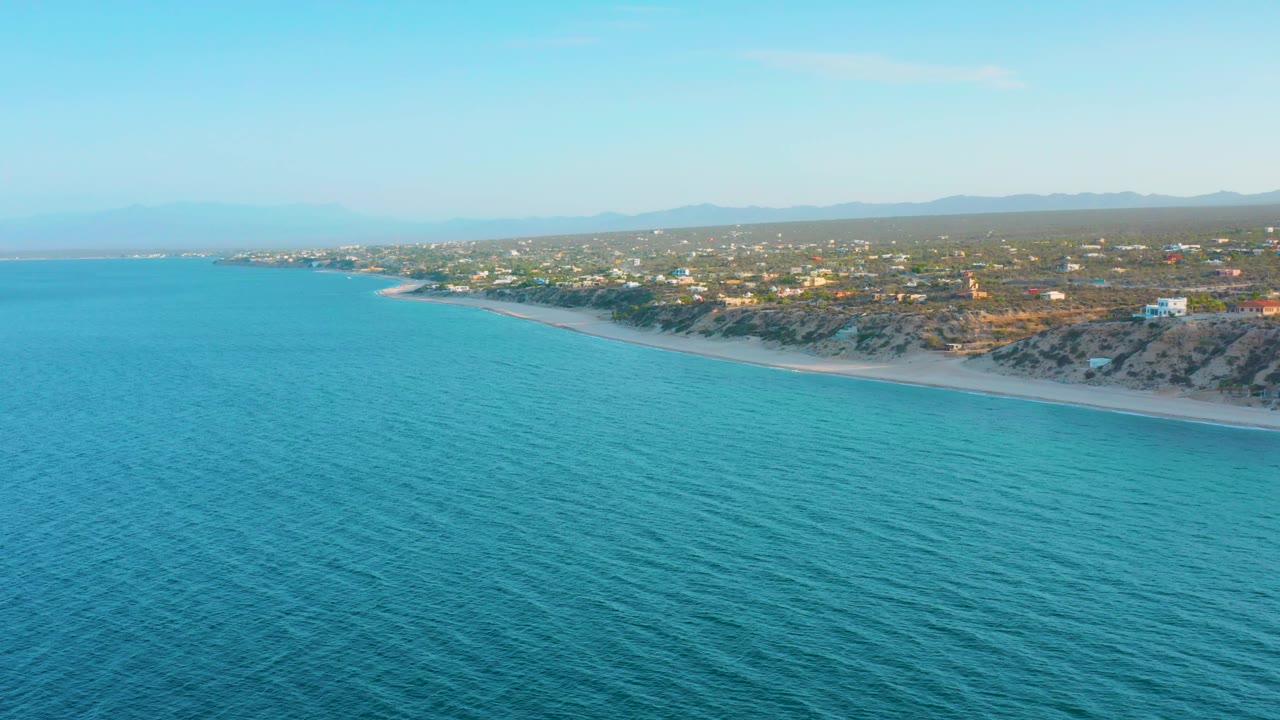 Wide drone-height view of the El Sargento La Ventana coast, with rippled turquoise water, a narrow beach strip, dry inland vegetation, scattered homes, and no visible people.