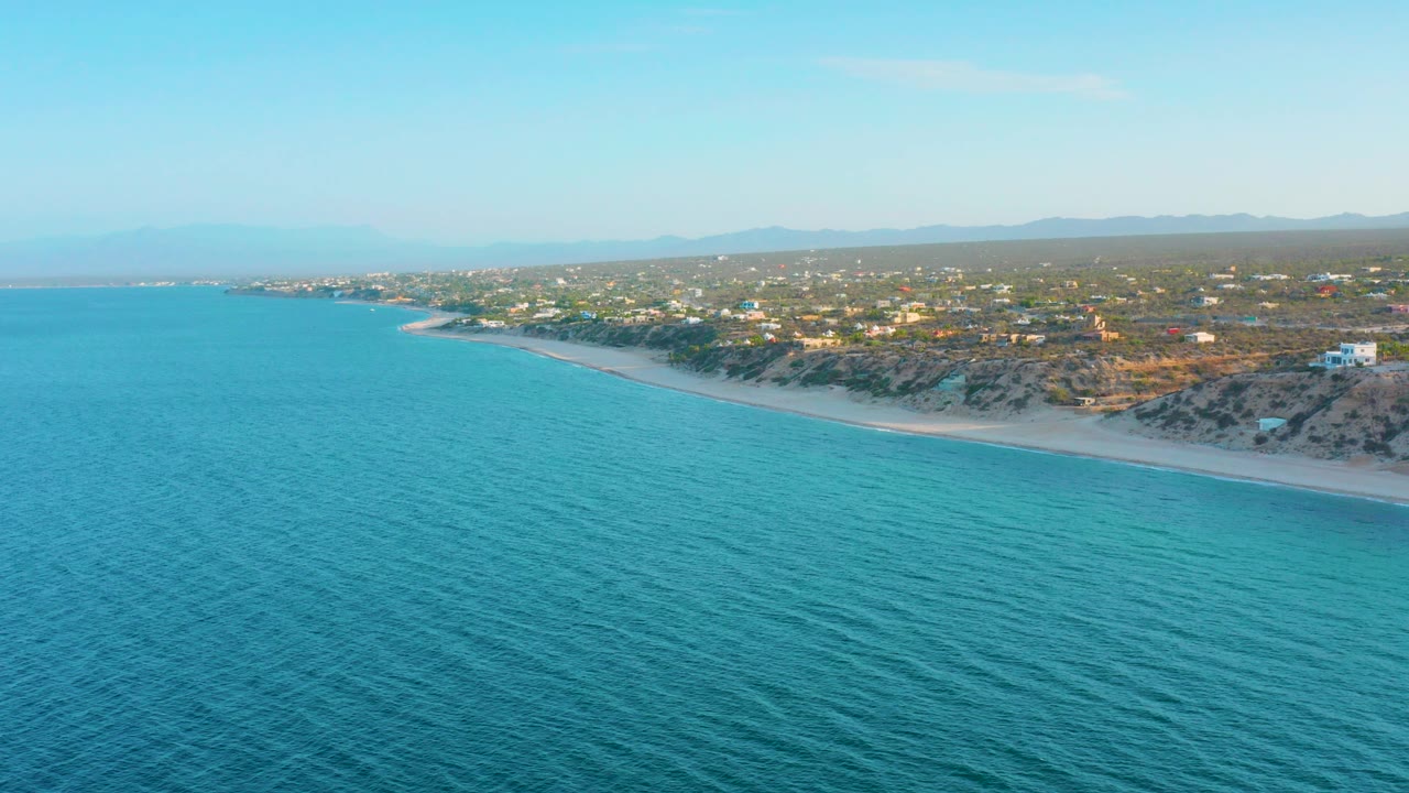 Aerial frame over blue-green water in El Sargento La Ventana, Mexico, with a pale beach, scattered coastal homes, scrub vegetation, distant hills, and no visible people.