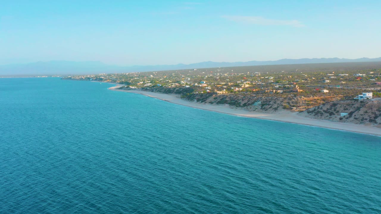 The aerial view continues along the El Sargento La Ventana shoreline, showing rippled turquoise water, a narrow beach, low vegetation, scattered homes, hazy hills, and no visible people.