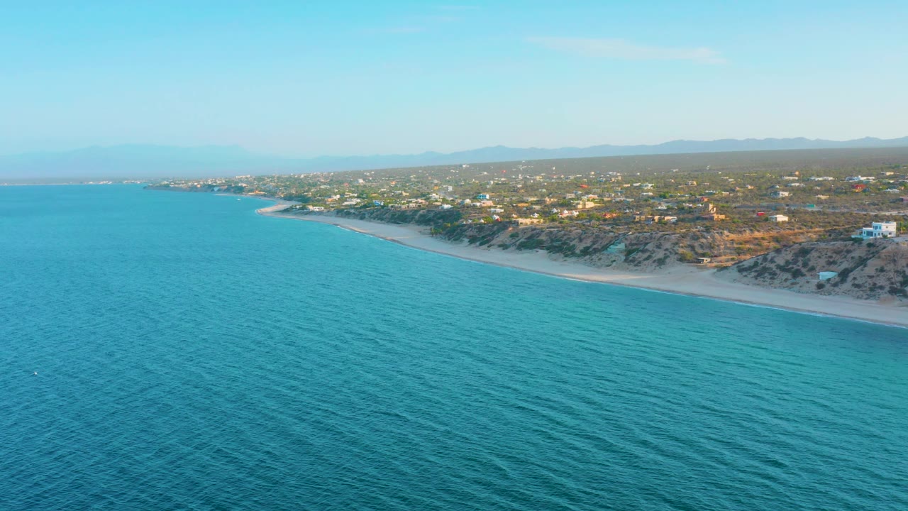 A wide aerial frame shows the El Sargento La Ventana coast with more open water in the foreground, a curved sandy beach, residential buildings inland, distant mountains, and no visible people.