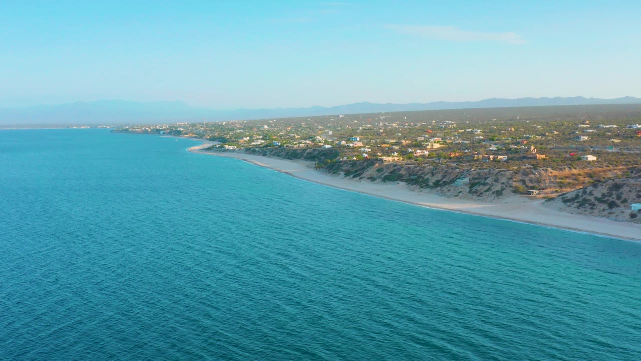 Wide aerial view over turquoise water toward a curving sandy shore and low buildings among green trees in El Sargento La Ventana, Mexico, with no visible people.