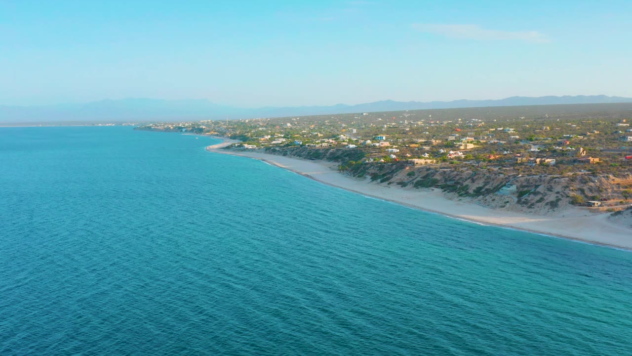 A second aerial frame holds over turquoise water with the sandy El Sargento La Ventana shoreline running diagonally below low dunes, scattered buildings, distant hills, and no visible people.