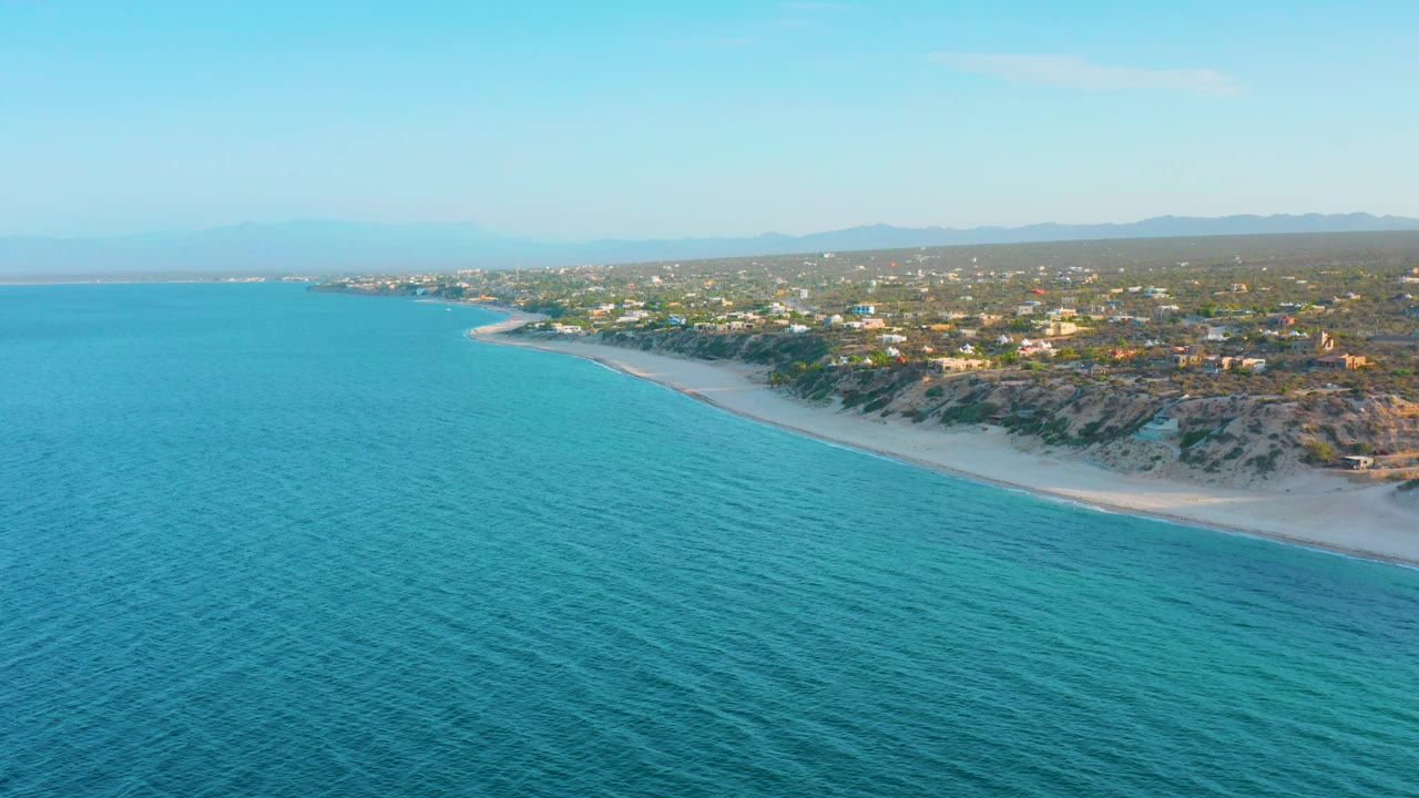 Wide drone frame looking across textured blue water to a pale beach backed by scrubby dunes, small buildings in El Sargento La Ventana, a hazy ridge line, and no visible people.