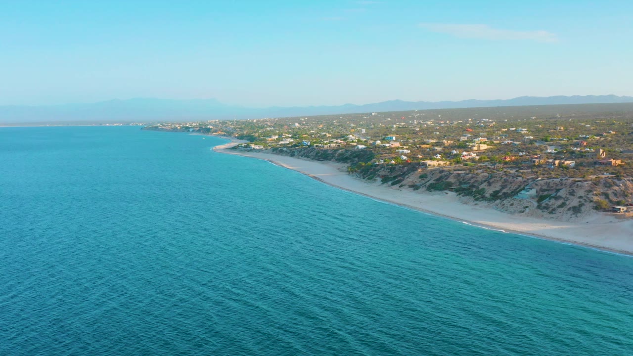 Aerial shoreline view with calm water, sandy beach, dunes, and low buildings.