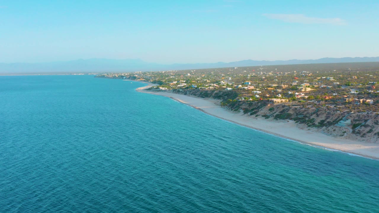 Aerial view over El Sargento La Ventana, Mexico, with turquoise sea filling the foreground, a pale beach curving along low coastal scrub, scattered buildings inland, hazy hills beyond, and no visible people.